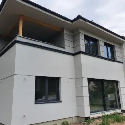 Nowoczesny dwupiętrowy dom z jasnoszarą elewacją, czarnymi ramami okiennymi i drewnianą konstrukcją balkonu, częściowo w stanie surowym z widoczną czerwoną cegłą na bocznej ścianie.