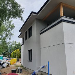 Kamil Popiel - Nowoczesny dom jednorodzinny z szarą elewacją w trakcie budowy, widoczny niezabezpieczony fragment balkonu z drewnianą konstrukcją, w tle żółty kontener na gruz i drzewa.