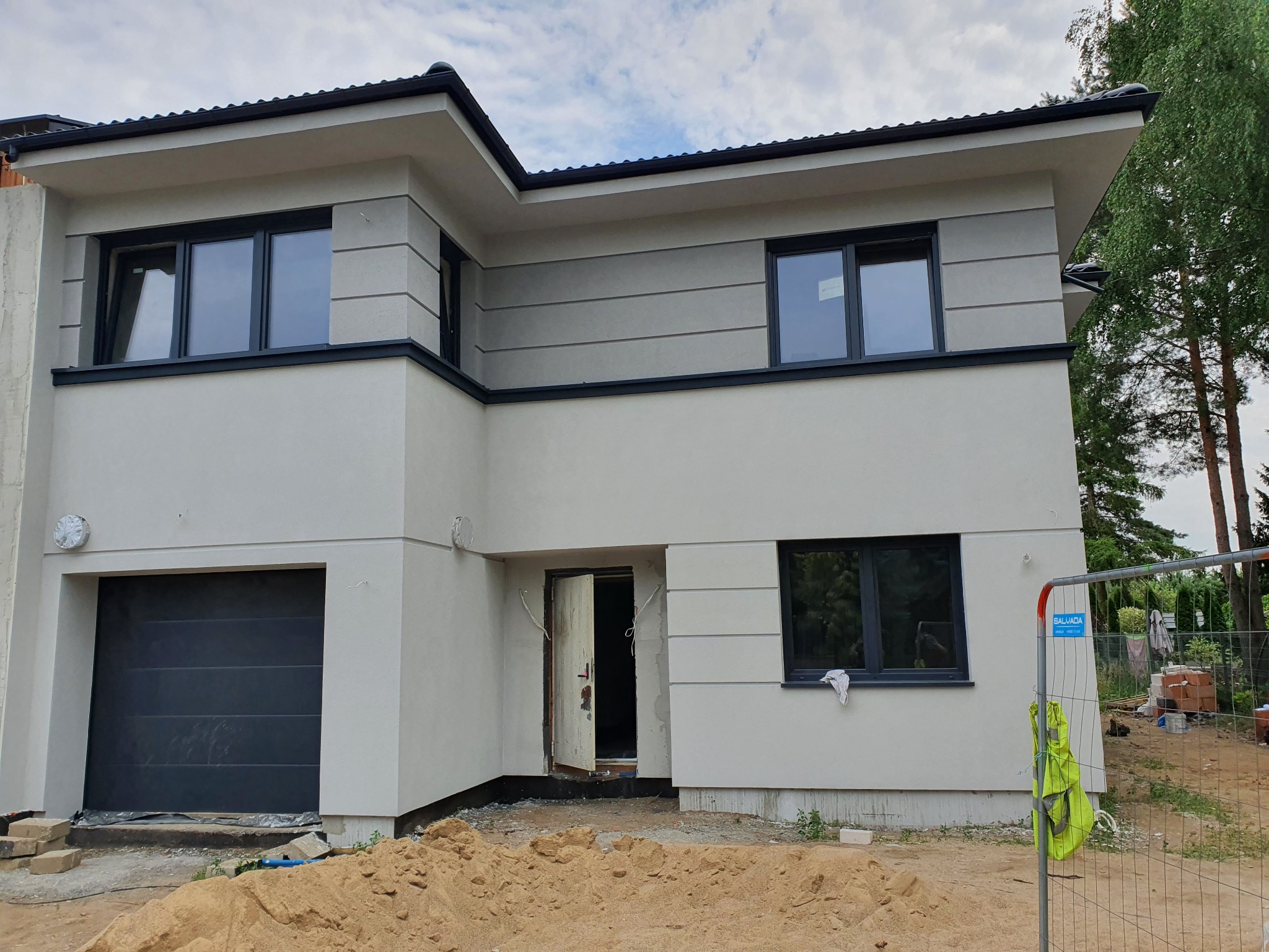 Dwukondygnacyjny dom jednorodzinny z płaskim dachem, wykończony jasnym tynkiem strukturalnym z ciemnymi ramami okiennymi i bramą garażową, widoczny piasek budowlany przed wejściem i tymczasowe...