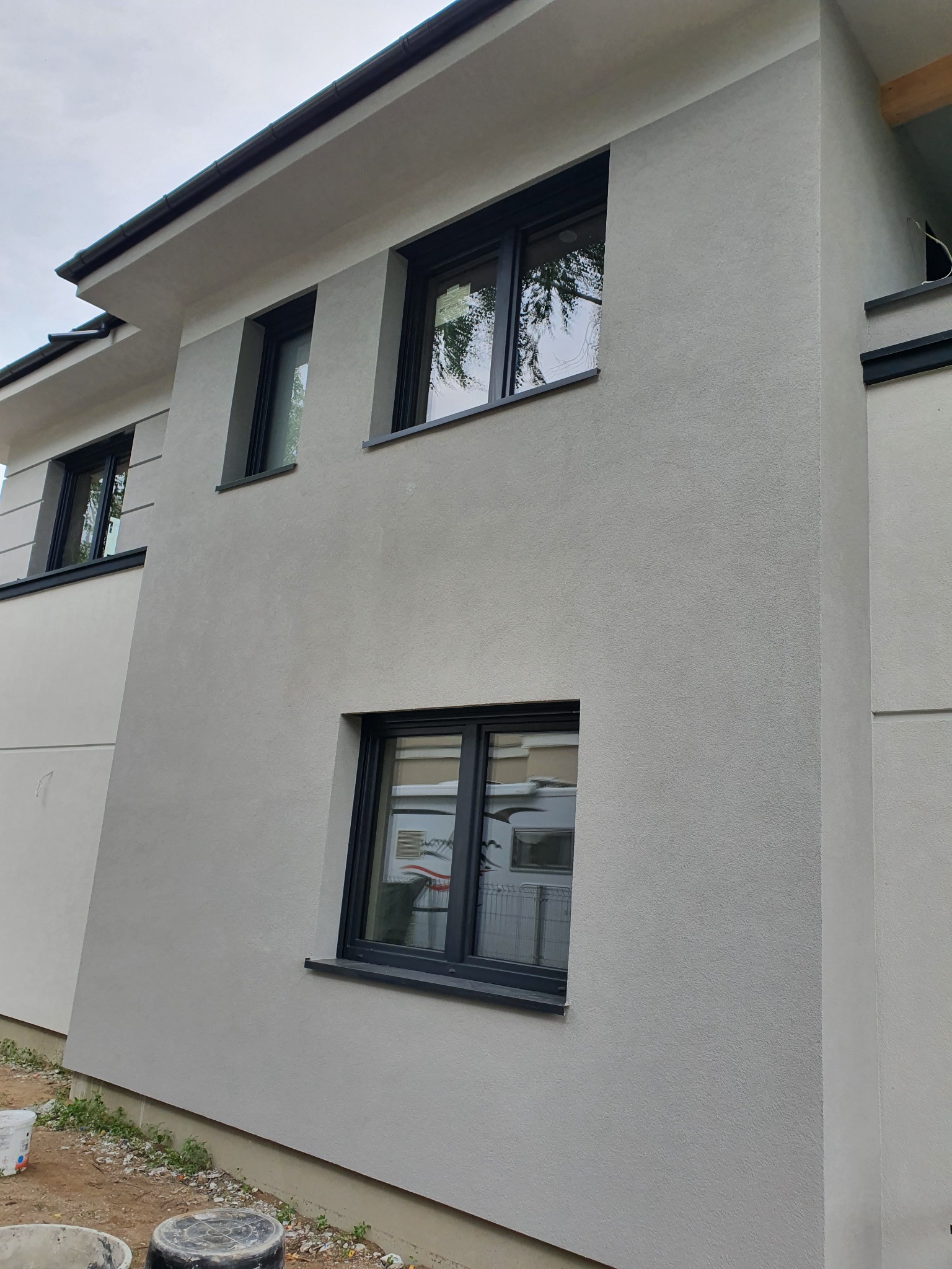 Nowoczesny dom jednorodzinny w Gorzowie Wielkopolskim z szarą elewacją o strukturze baranka, czarne ramy okienne i widoczne wiaderka po farbie na dole kadru.