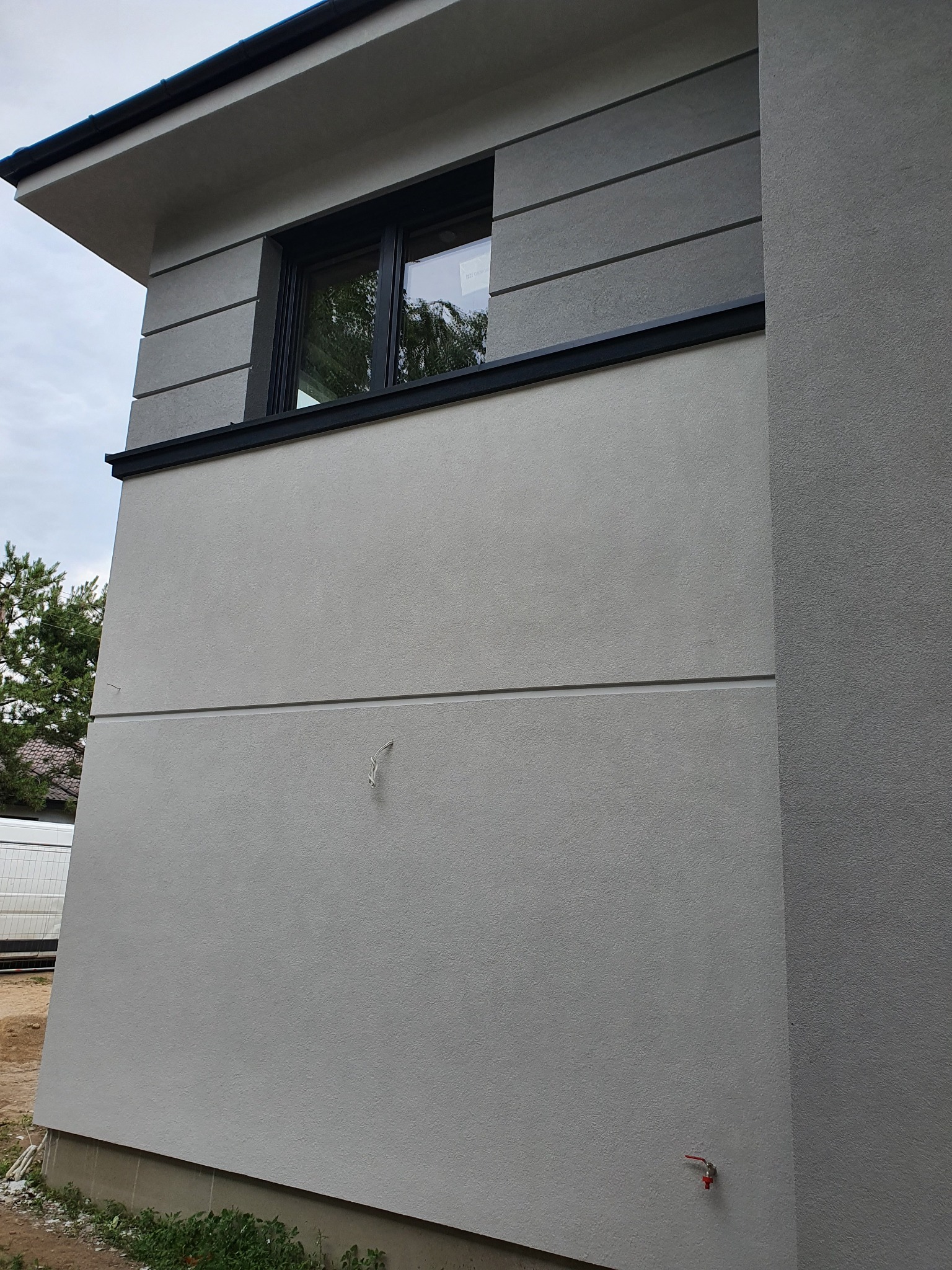 Fragment ściany budynku po nałożeniu jasnoszarego tynku strukturalnego, z widocznym oknem w czarnej ramie i wystającym kranikiem przy ziemi. Widoczne przewody elektryczne wystające ze ściany.