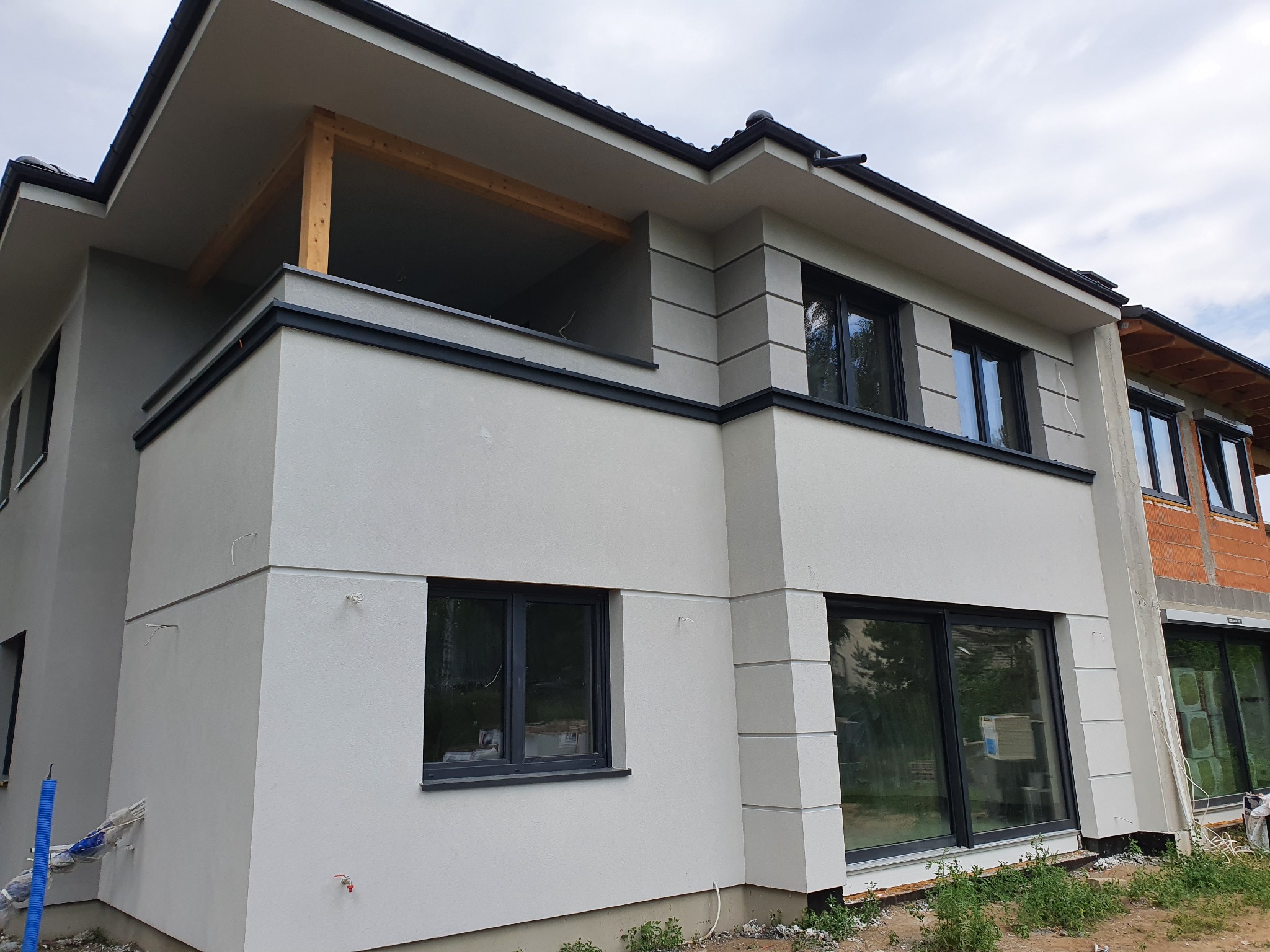 Nowoczesny dwupiętrowy dom z jasnoszarą elewacją, czarnymi ramami okiennymi i drewnianą konstrukcją balkonu, częściowo w stanie surowym z widoczną czerwoną cegłą na bocznej ścianie.