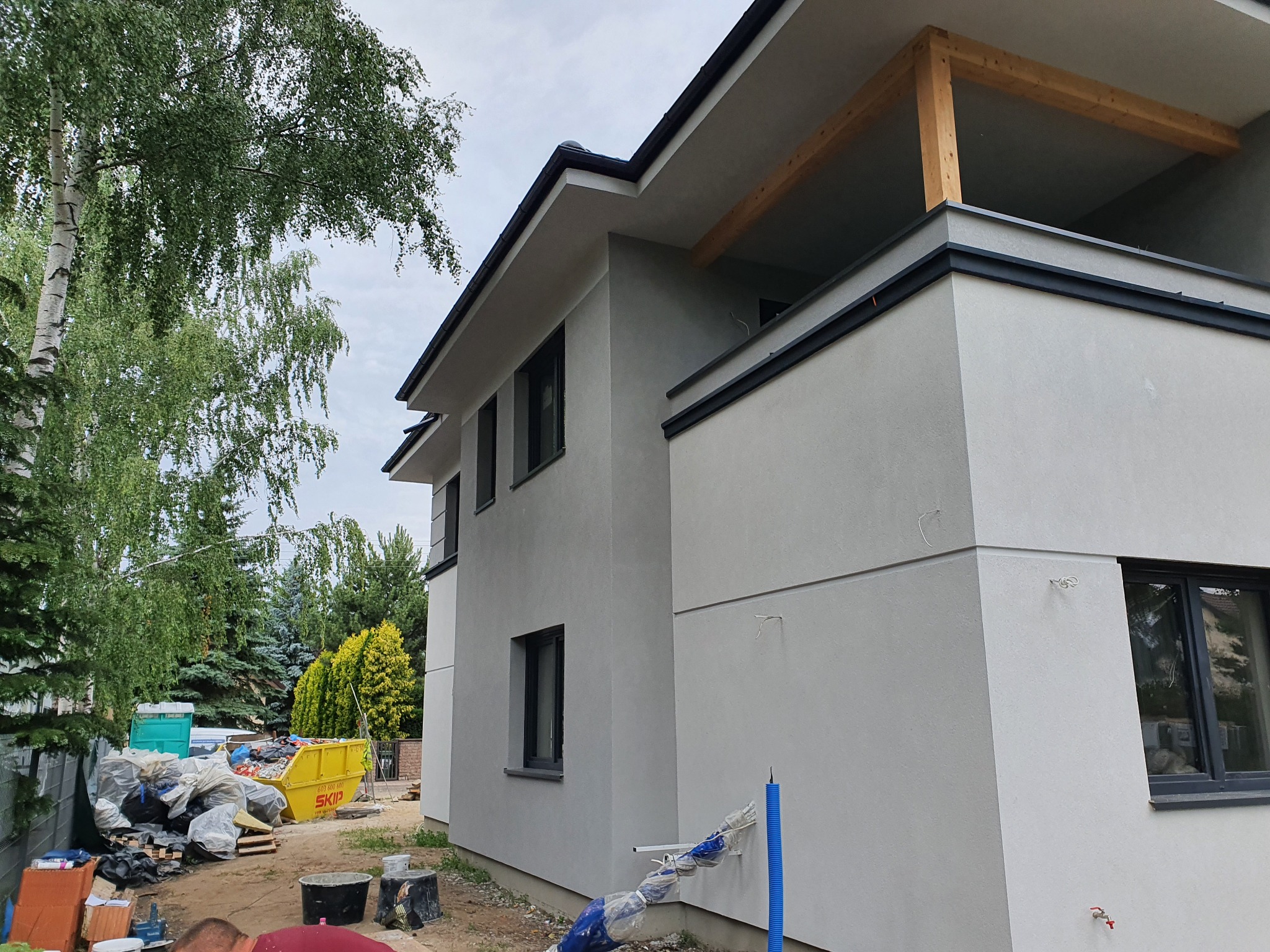 Nowoczesny dom jednorodzinny z szarą elewacją w trakcie budowy, widoczny niezabezpieczony fragment balkonu z drewnianą konstrukcją, w tle żółty kontener na gruz i drzewa.