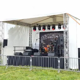 Scena plenerowa z metalową barierką, nagłośnieniem po bokach, perkusją i muzykiem w skórzanej kurtce przed banerem zespołu Corrupha.
