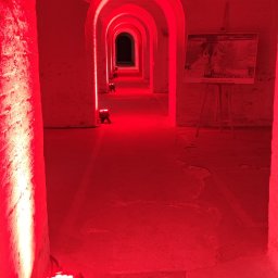 MG Acoustics Mikołaj Grzechociński - Czerwone, tunelowe przejście w Warszawie, oświetlone przez lampy LED. Na końcu tunelu widoczna ciemność, a po prawej stronie stoi sztaluga z obrazem.