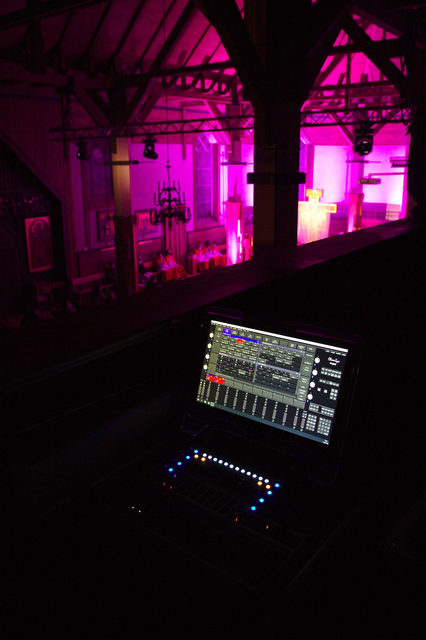 Panel sterowania konsolety oświetleniowej i nagłośnieniowej z widokiem na salę oświetloną na różowo, z elementami drewnianej konstrukcji dachu.