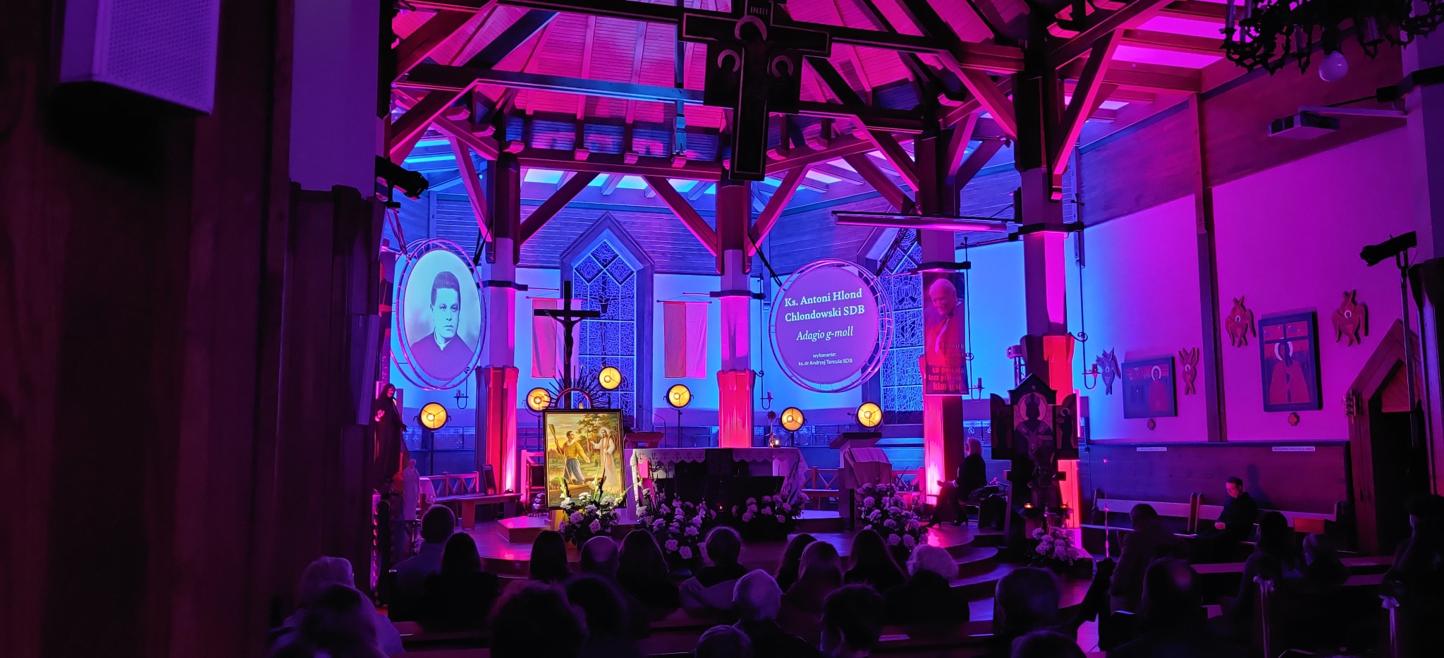 Wnętrze kościoła oświetlone niebieskim i różowym światłem, z portretami i dekoracjami, przygotowane na uroczystość, widoczni ludzie w ławkach.