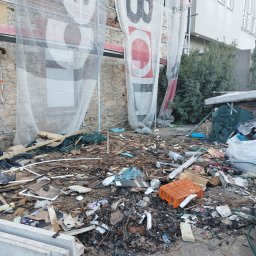 Usługi Sprzątające MKG - Opróżnianie terenu budowy: gruz, odpady budowlane i resztki materiałów pod rusztowaniem z reklamą. Widoczne elementy starej kamiennej ściany i fragment budynku.