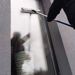 Usługi Sprzątające MKG - Osoba myje okno z zewnątrz budynku za pomocą mopa na kiju. Na szybie widoczne smugi wody i odbicie otoczenia.