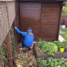 Usługi Sprzątające MKG - Mężczyzna maluje drewniany domek ogrodowy na brązowo, siedząc obok rabatki z ziołami i truskawkami. Widoczny fragment pomalowanego płotu.