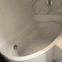 Biała, narożna wanna z armaturą i mozaiką na ścianach. Widok z góry prezentuje czystą, gotową do użycia łazienkę z elementami dekoracyjnymi.