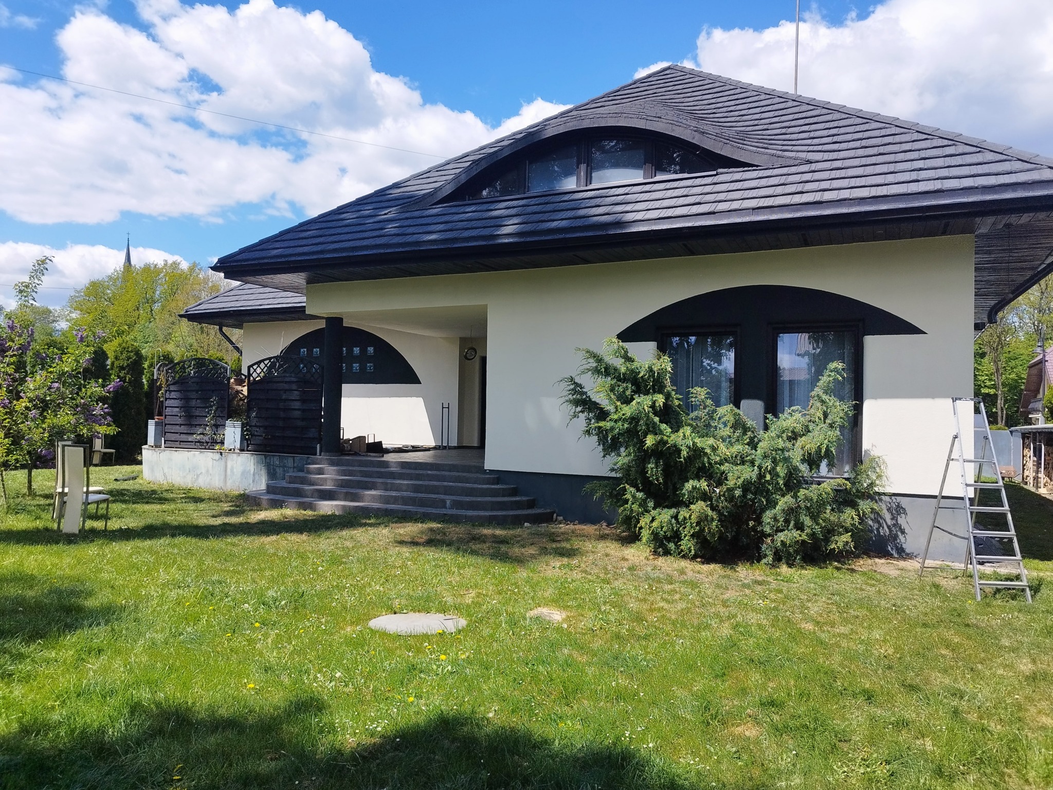 Dom jednorodzinny z ciemną dachówką i ozdobnymi oknami na poddaszu, zielony trawnik, drabina oparta o ścianę. Słoneczny dzień.