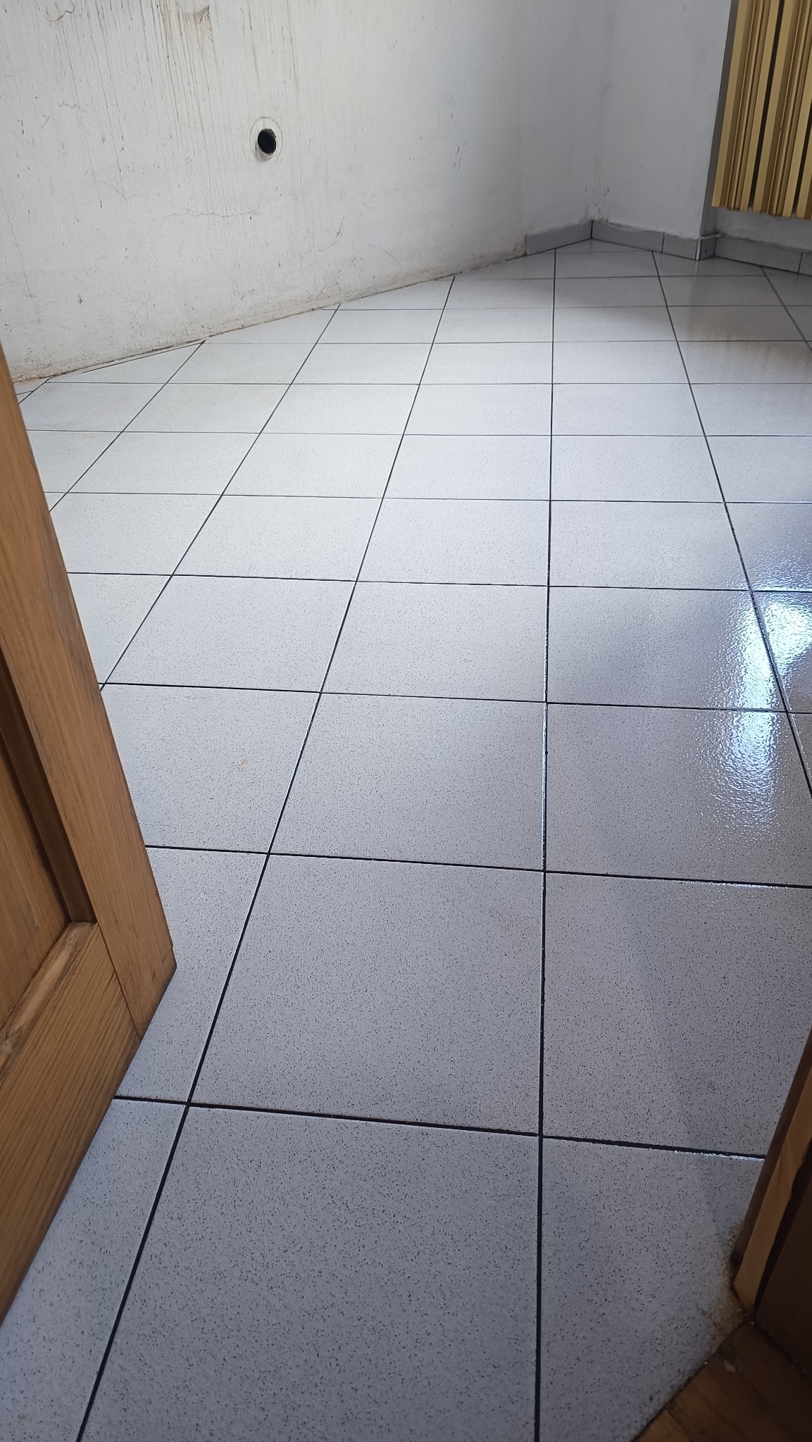 Białe, kwadratowe płytki podłogowe po umyciu, widoczne od otwartych drzwi, z refleksami światła i czystymi fugami. Ściana biała, w rogu widoczne zasłony.