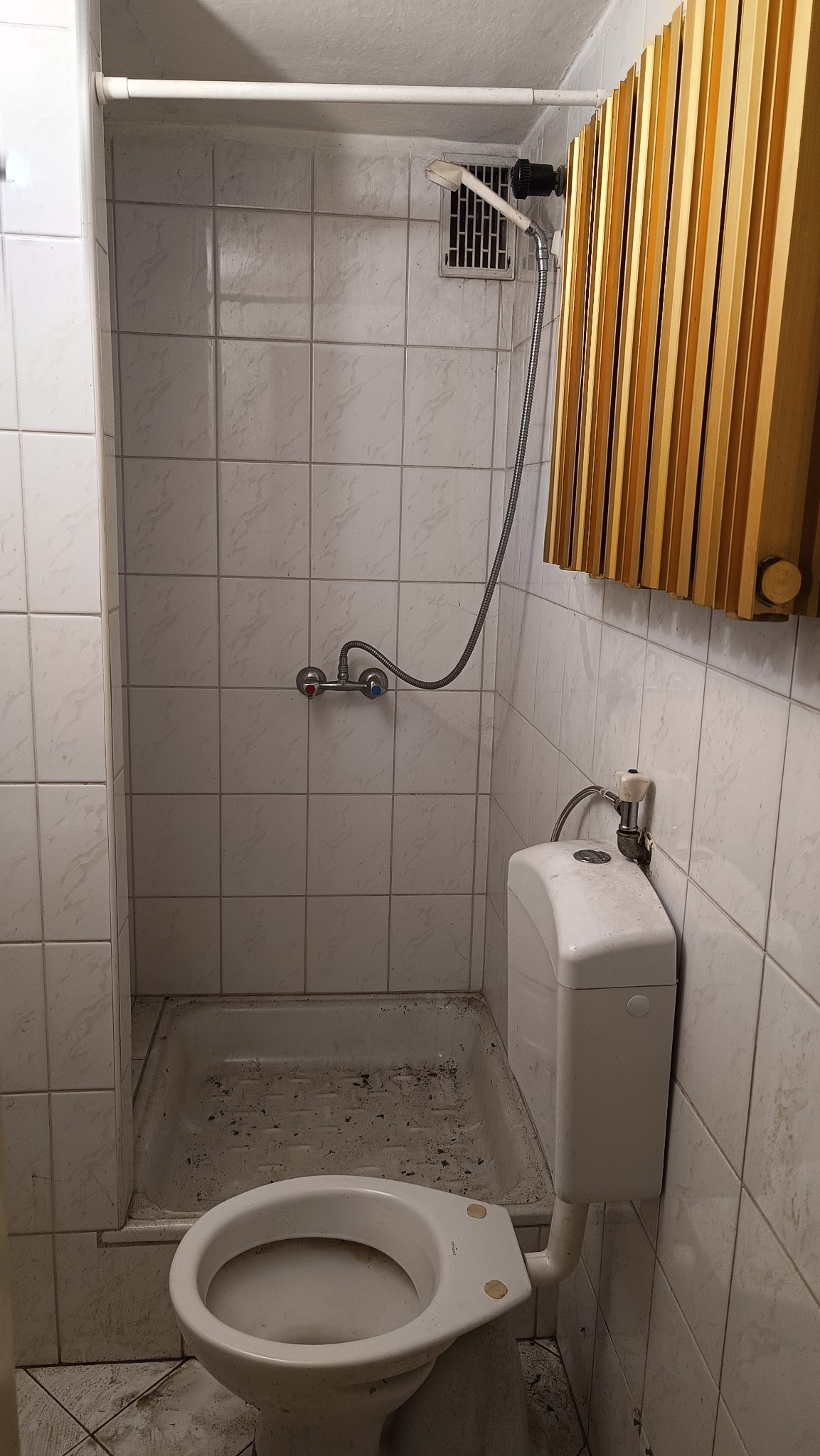 Brudna toaleta z kabiną prysznicową w łazience wyłożonej kwadratowymi płytkami. Widoczny grzejnik. Zaniedbany stan.