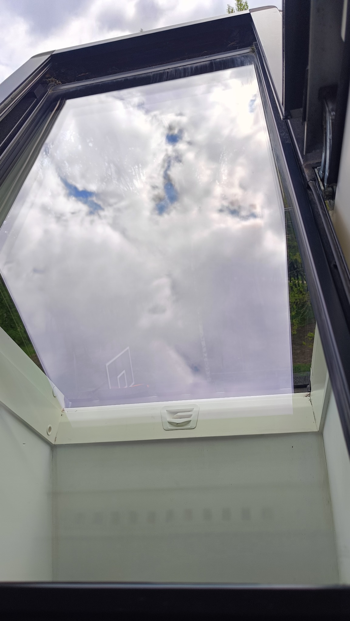 Uchylone okno dachowe po umyciu, z widocznym odbiciem chmur. Czysta szyba i biała rama. Widok z dołu, brak smug i zacieków.