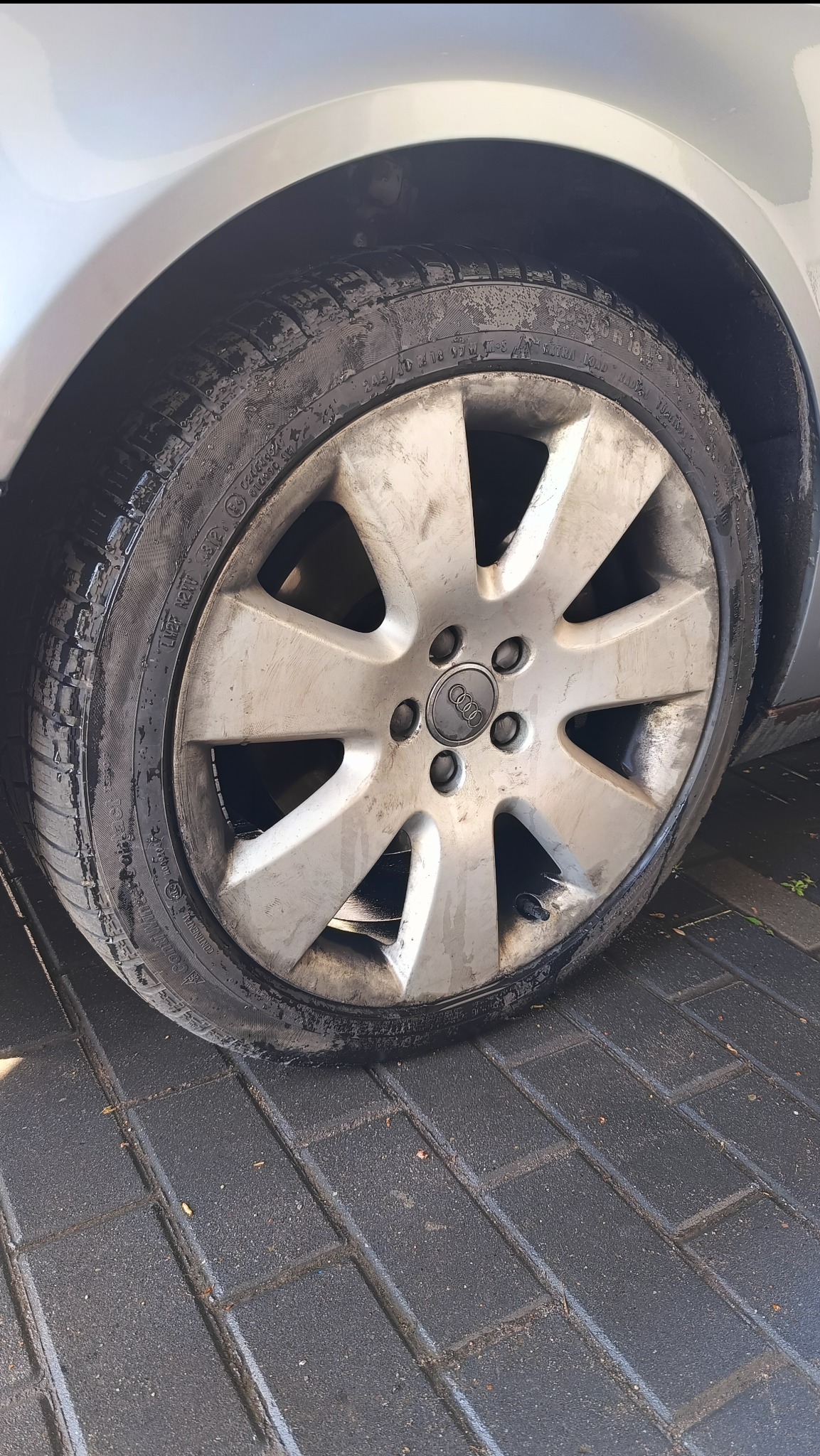 Brudne koło aluminiowe Audi z wyraźnym bieżnikiem opony, stojące na mokrej kostce brukowej. Widoczne zabrudzenia na feldze i oponie.