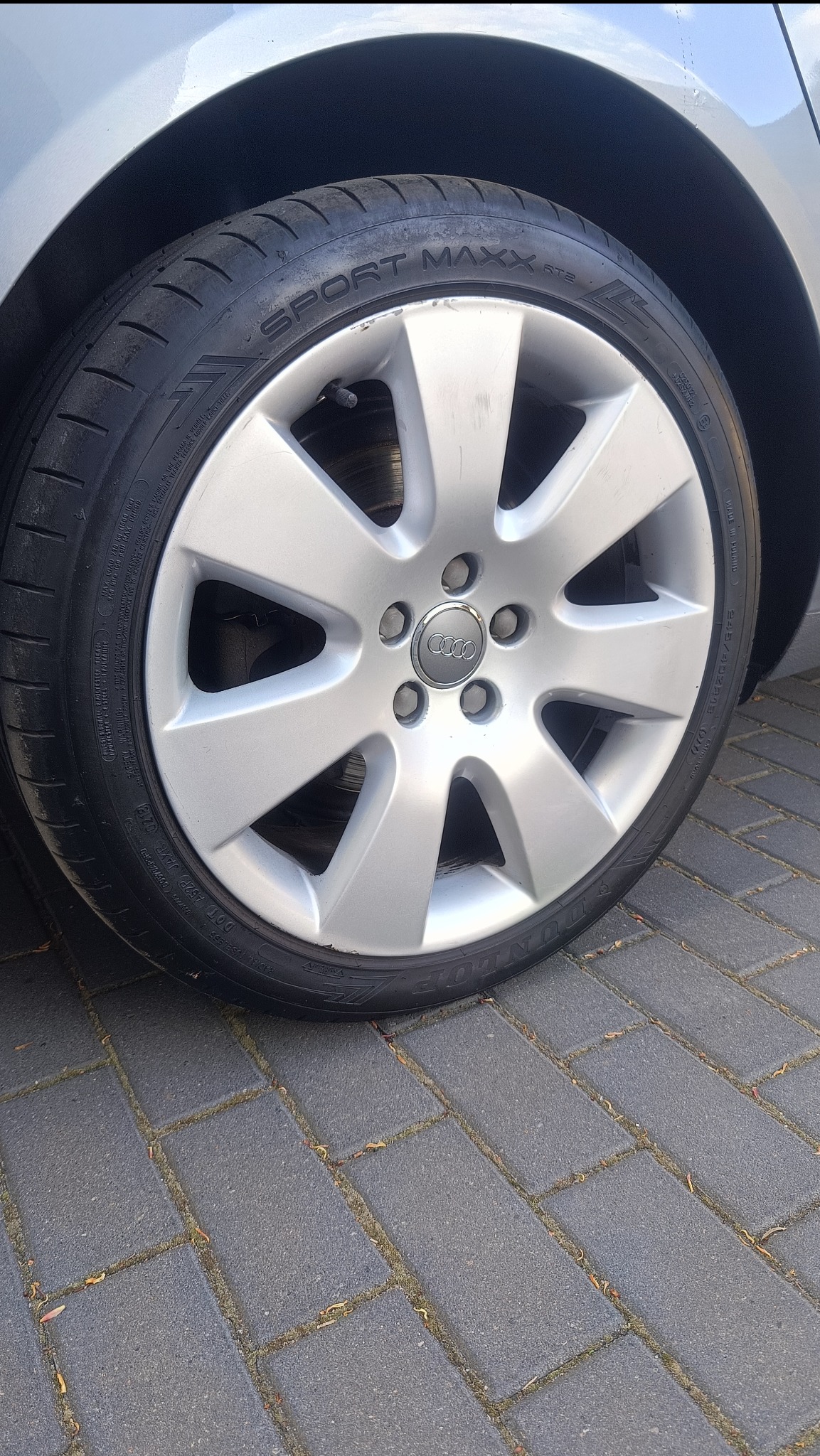 Błyszcząca felga Audi Sport Maxx na tle kostki brukowej. Widoczne detale opony i srebrna felga z logo Audi. Kąt ujęcia podkreśla nowoczesny design.