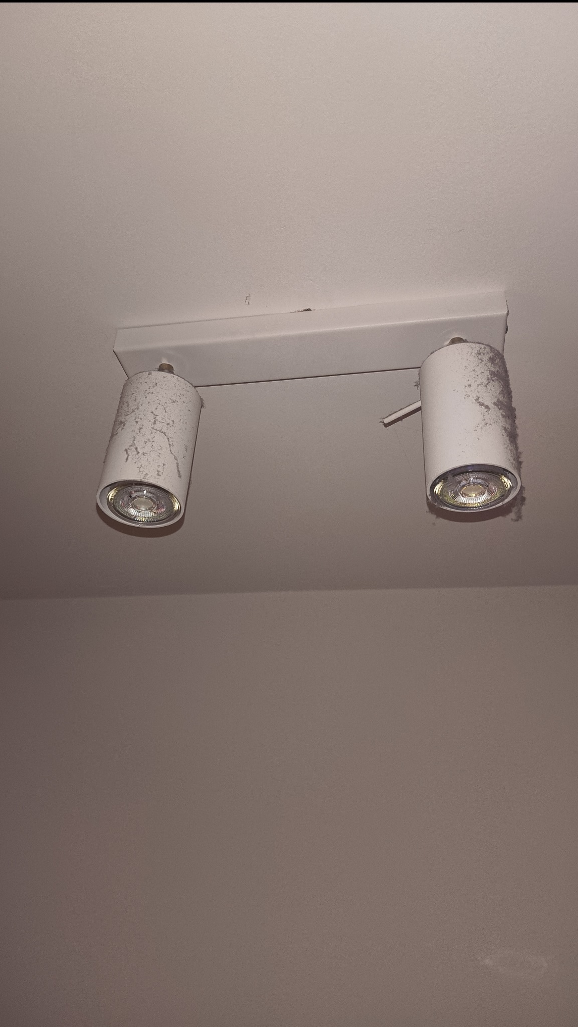 Biała lampa sufitowa z dwoma reflektorami, pokryta grubą warstwą kurzu, zamontowana na białym suficie. Widoczne żarówki.