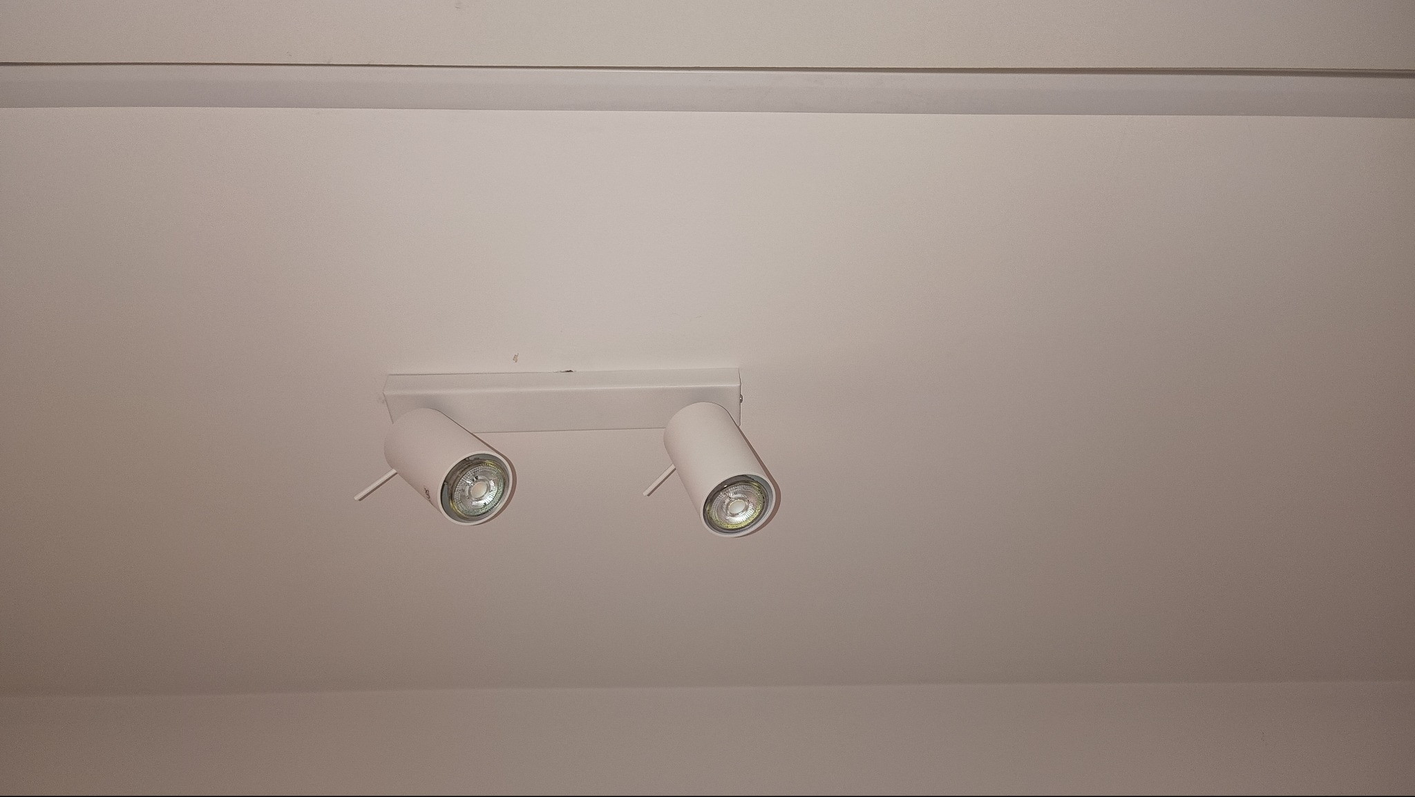 Biała, podwójna lampa sufitowa typu reflektor z żarówkami LED, zamontowana na gładkim, jasnym suficie. Minimalistyczny design oświetlenia.