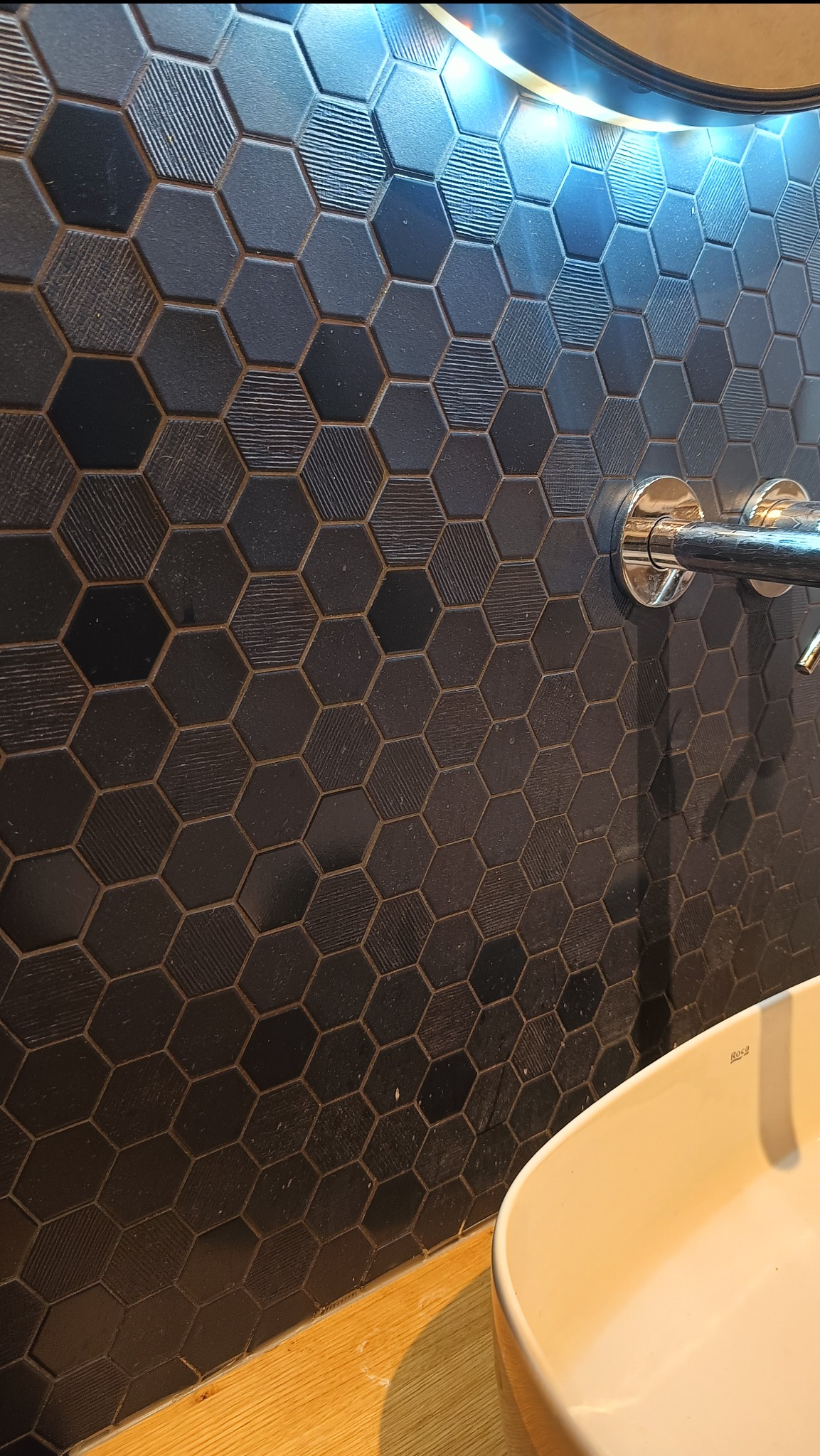 Ściana wyłożona heksagonalnymi, czarnymi płytkami z różnymi fakturami, oświetlona od góry, z fragmentem umywalki i baterii. Wnętrze łazienki.