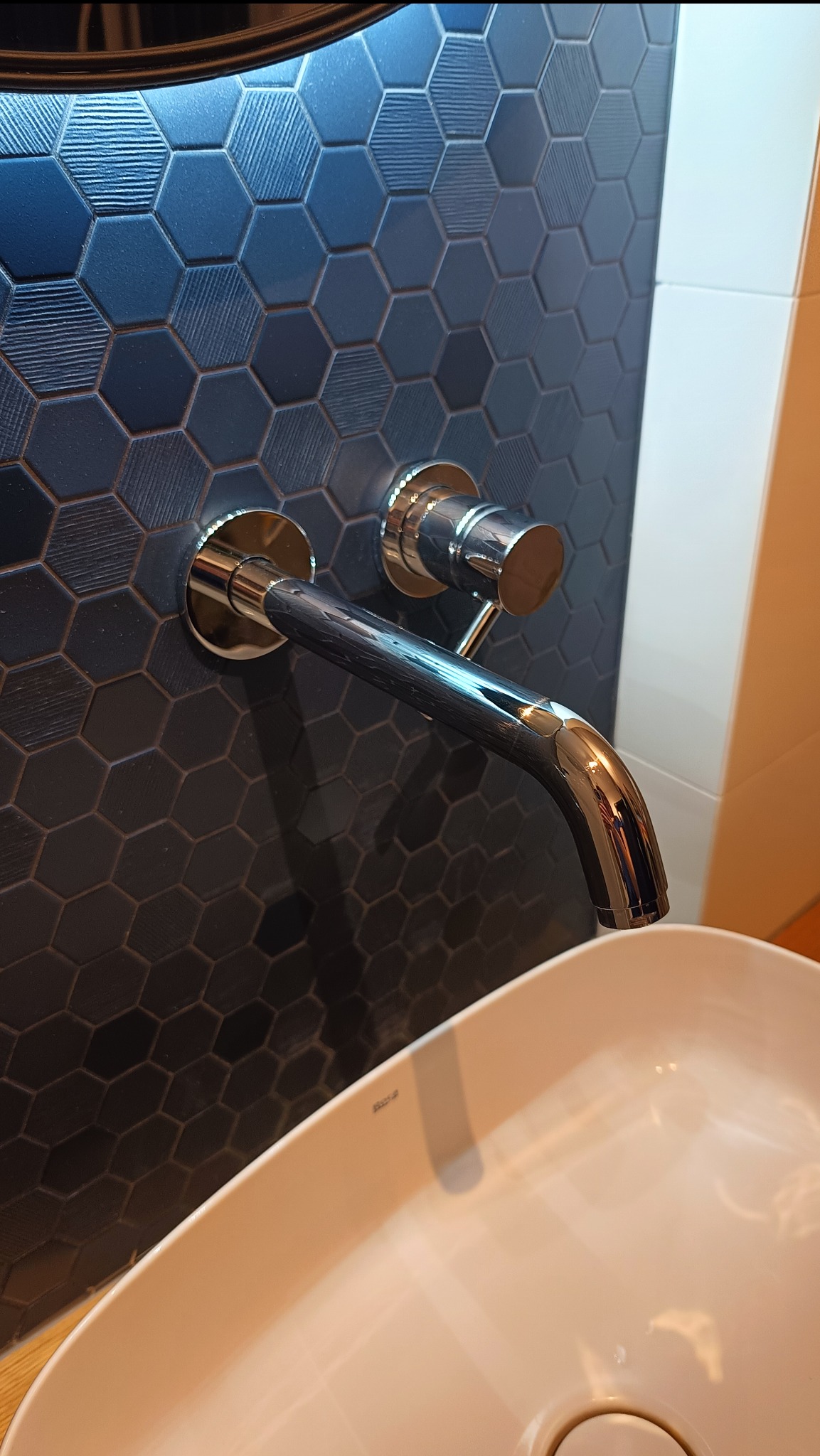 Bateria umywalkowa chromowana na tle ściany z ciemnych, heksagonalnych płytek. Umywalka ceramiczna o zaokrąglonym kształcie, nowoczesny design.