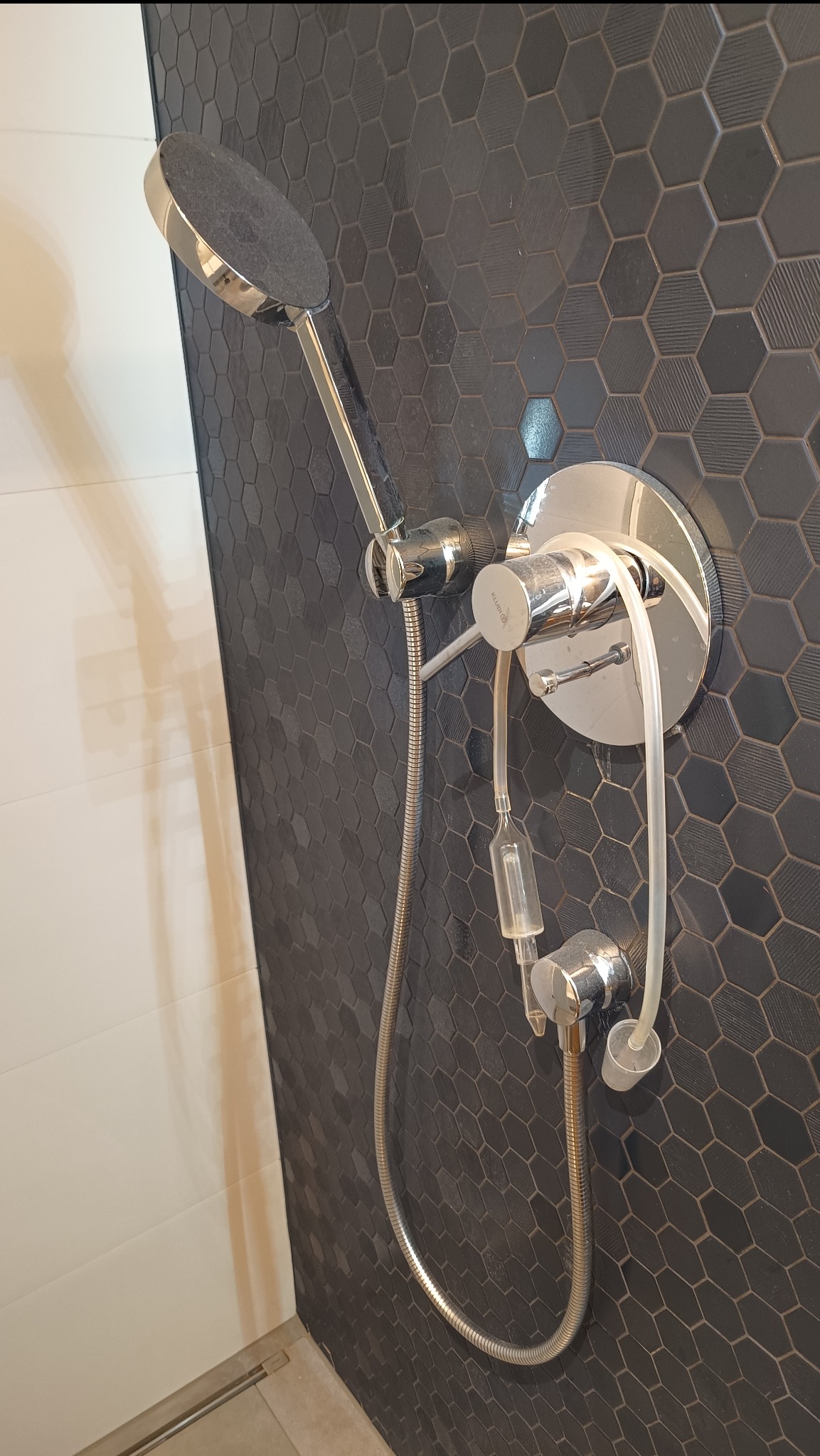 Chromowana bateria prysznicowa z deszczownicą na tle ściany z heksagonalnych, grafitowych płytek. Widoczny biały panel boczny i fragment odpływu liniowego.
