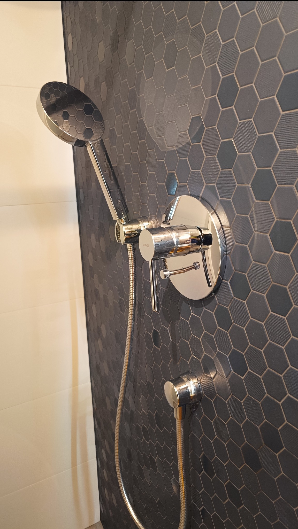 Bateria prysznicowa z deszczownicą na ścianie wyłożonej ciemnymi, heksagonalnymi płytkami. Widoczny chromowany panel sterujący i wąż doprowadzający wodę.
