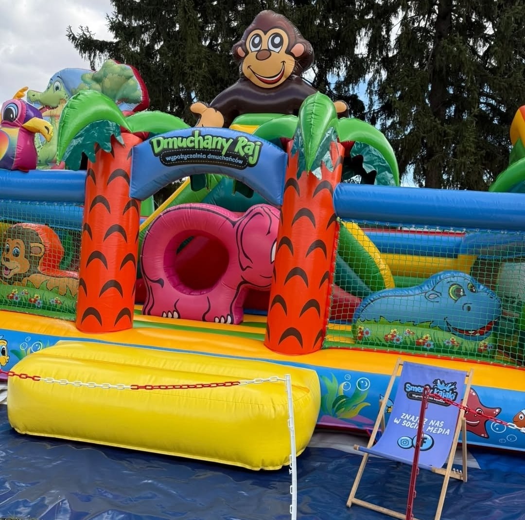 Kolorowy dmuchany plac zabaw z motywami zwierzęcymi, w tym małpa na szczycie i napis 'Dmuchany Raj'. W tle drzewa i krzesło plażowe z logo firmy.