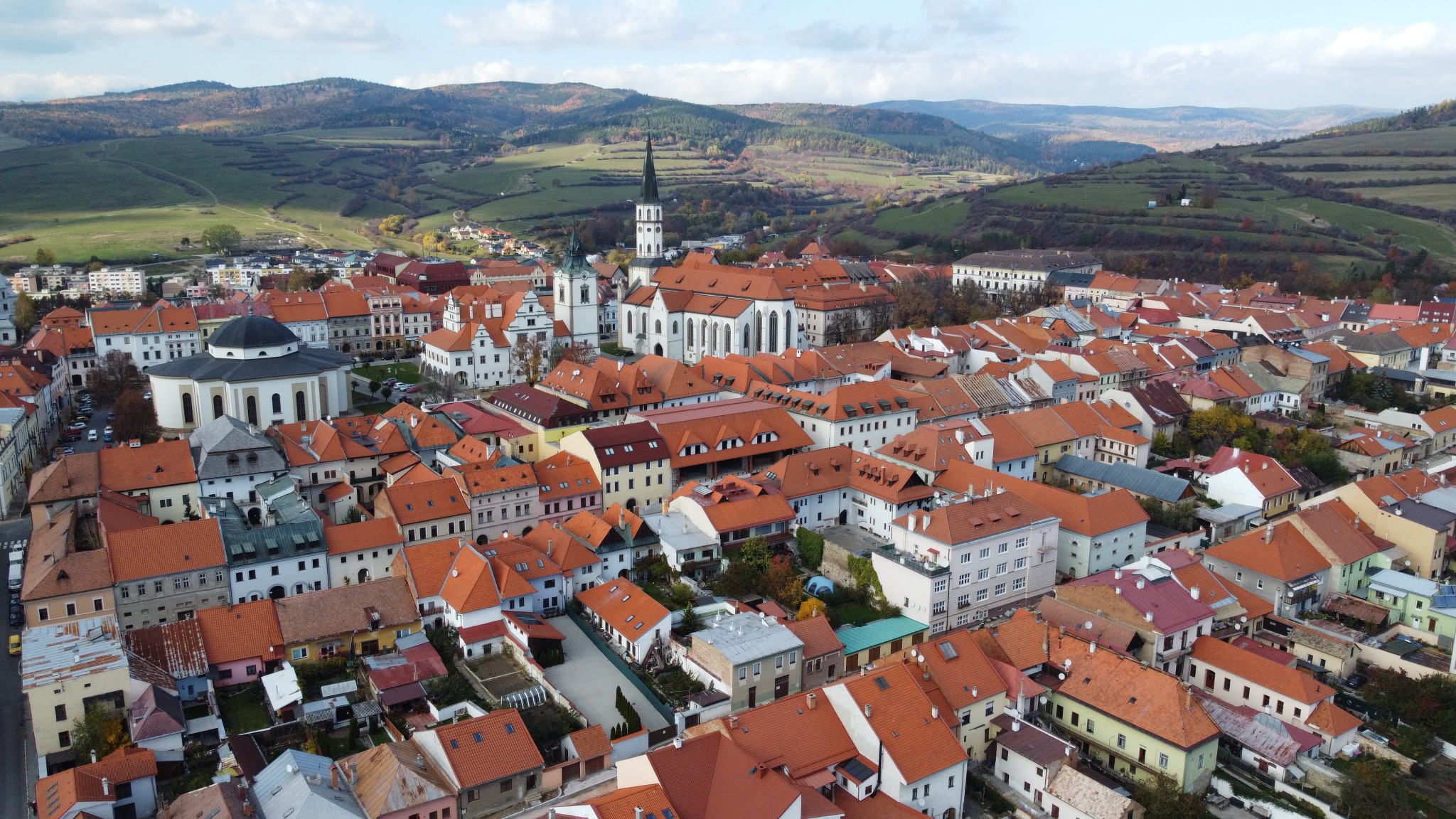 Panoramiczny widok z lotu ptaka na miasteczko z czerwonymi dachami, kościołem z wysoką wieżą i pagórkowatym krajobrazem w tle.