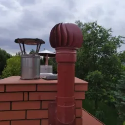 Czerwony komin z nasadą turbinową na ceglanym kominie, obok metalowy wkład kominowy z daszkiem, widok na zieleń i pochmurne niebo.