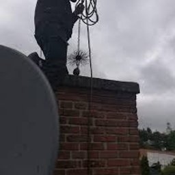 Usługi kominiarsko-gazownicze Paweł Danielik - Mężczyzna w czarnym ubraniu na dachu, trzyma linę z szczotką kominiarską nad kominem z czerwonej cegły, pochmurne niebo w tle.