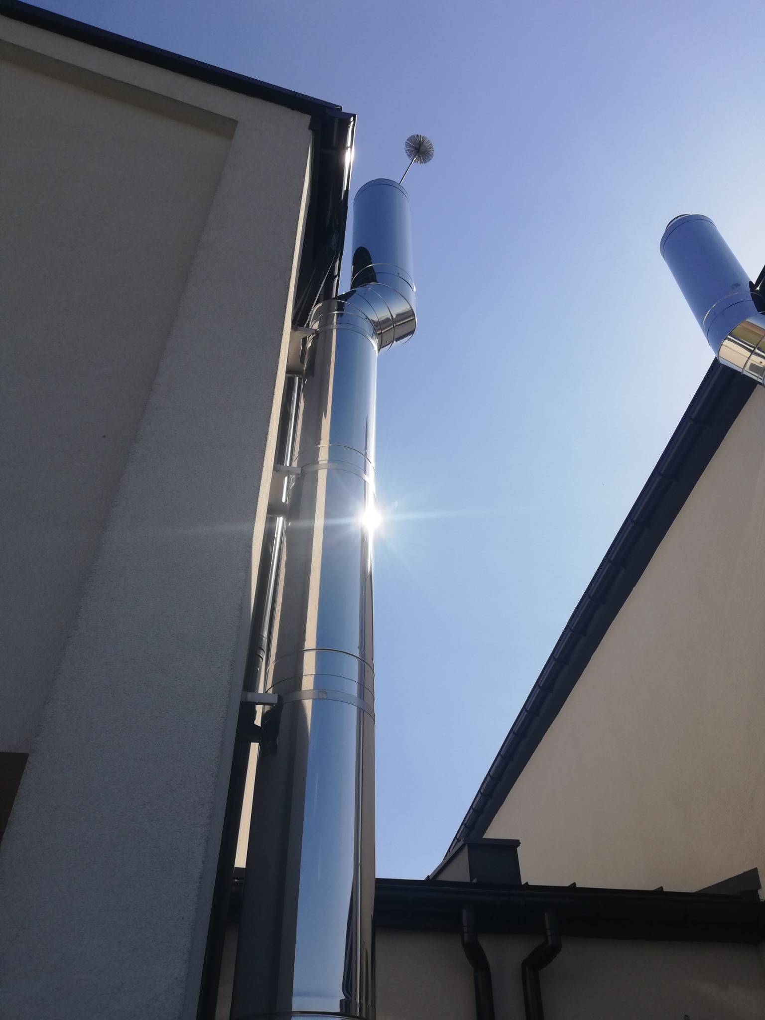 Dwa błyszczące, metalowe kominy zamontowane na ścianach budynków, widoczne z dołu pod kątem, jeden z zabezpieczeniem na szczycie, odbijające promienie słoneczne na tle błękitnego nieba.