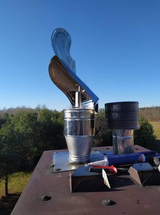 Instalacja metalowego wkładu kominowego z daszkiem na dachu, widoczne narzędzia i uszczelniacz, w tle drzewa i błękitne niebo.