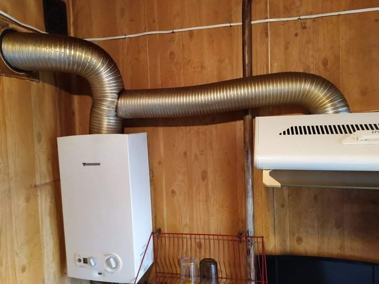 Biały piec gazowy z podłączonym, elastycznym przewodem wentylacyjnym w pomieszczeniu wykończonym boazerią; widoczny fragment okapu kuchennego i półka z naczyniami.