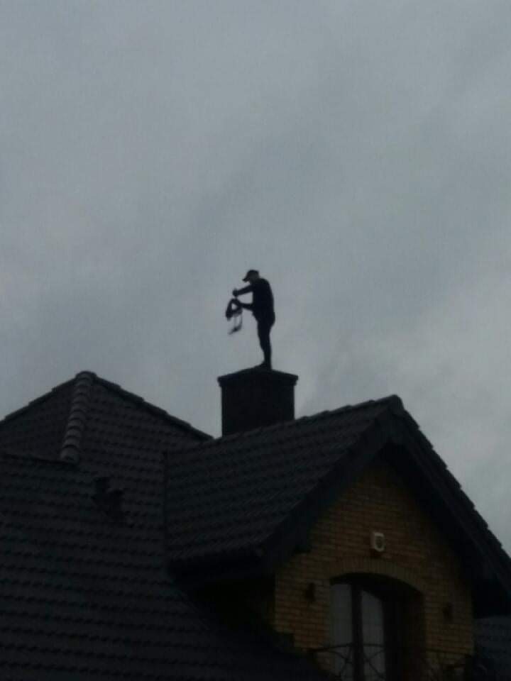 Sylwetka osoby na kominie domu, trzymającej narzędzia, na tle pochmurnego nieba.