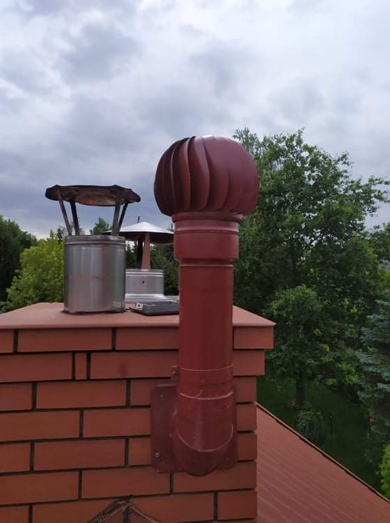 Czerwony komin z nasadą turbinową na ceglanym kominie, obok metalowy wkład kominowy z daszkiem, widok na zieleń i pochmurne niebo.