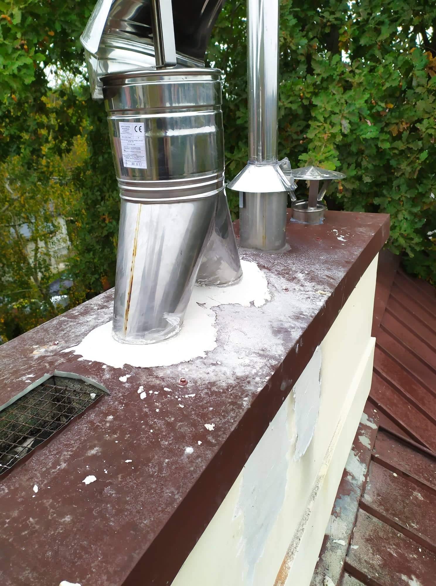 Metalowy komin z uszczelnieniem na dachu, widoczne ślady korozji na elemencie komina i zabrudzenia na dachu