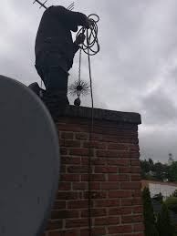 Mężczyzna w czarnym ubraniu na dachu, trzyma linę z szczotką kominiarską nad kominem z czerwonej cegły, pochmurne niebo w tle.