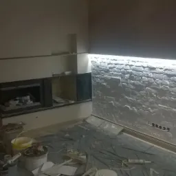 Ściana wykończona dekoracyjną cegłą z podświetleniem LED, widoczne narzędzia i materiały malarskie na podłodze zabezpieczonej folią, w tle fragment kominka.