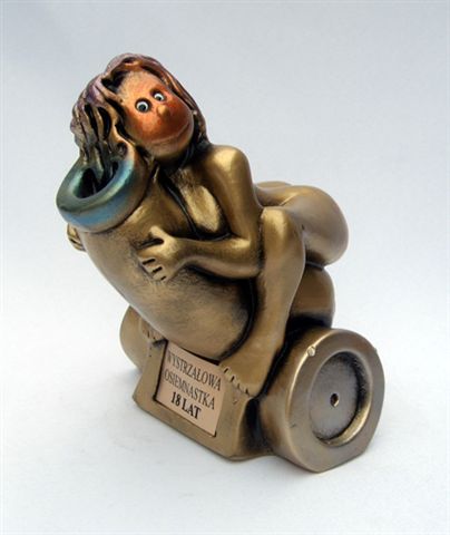 Figurka gipsowa przedstawiająca kobietę siedzącą na stylizowanej armacie, z napisem 'Wystrzałowa Osiemnastka 18 lat', na białym tle.