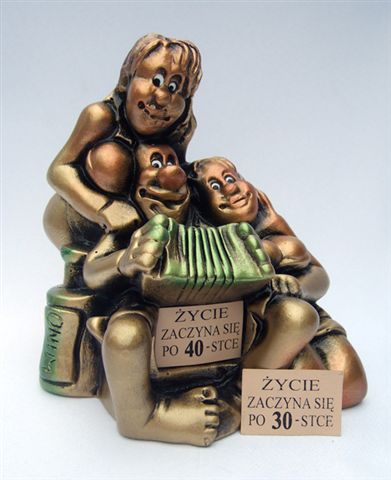 Gipsowa figurka przedstawiająca trzy postacie: kobietę obejmującą dwóch mężczyzn grających na akordeonie, z kartkami 'Życie zaczyna się po 40-stce' i 'Życie zaczyna się po 30-stce'.