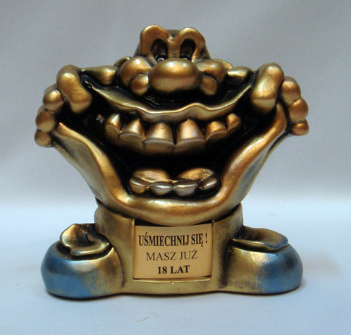 Figurka gipsowa przedstawiająca uśmiechniętą postać z napisem 'Uśmiechnij się! Masz już 18 lat', pomalowana na złoto z niebieskimi akcentami u podstawy.