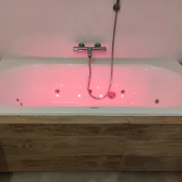 Biała wanna z hydromasażem podświetlona na czerwono, obudowana drewnopodobną okładziną, chromowana bateria i słuchawka prysznicowa, białe kafelki w tle.