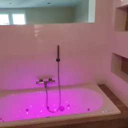 Biała wanna z hydromasażem podświetlona na różowo, z baterią prysznicową na ścianie z białych płytek, w tle lustro i wnęka z elementami drewnopodobnymi.