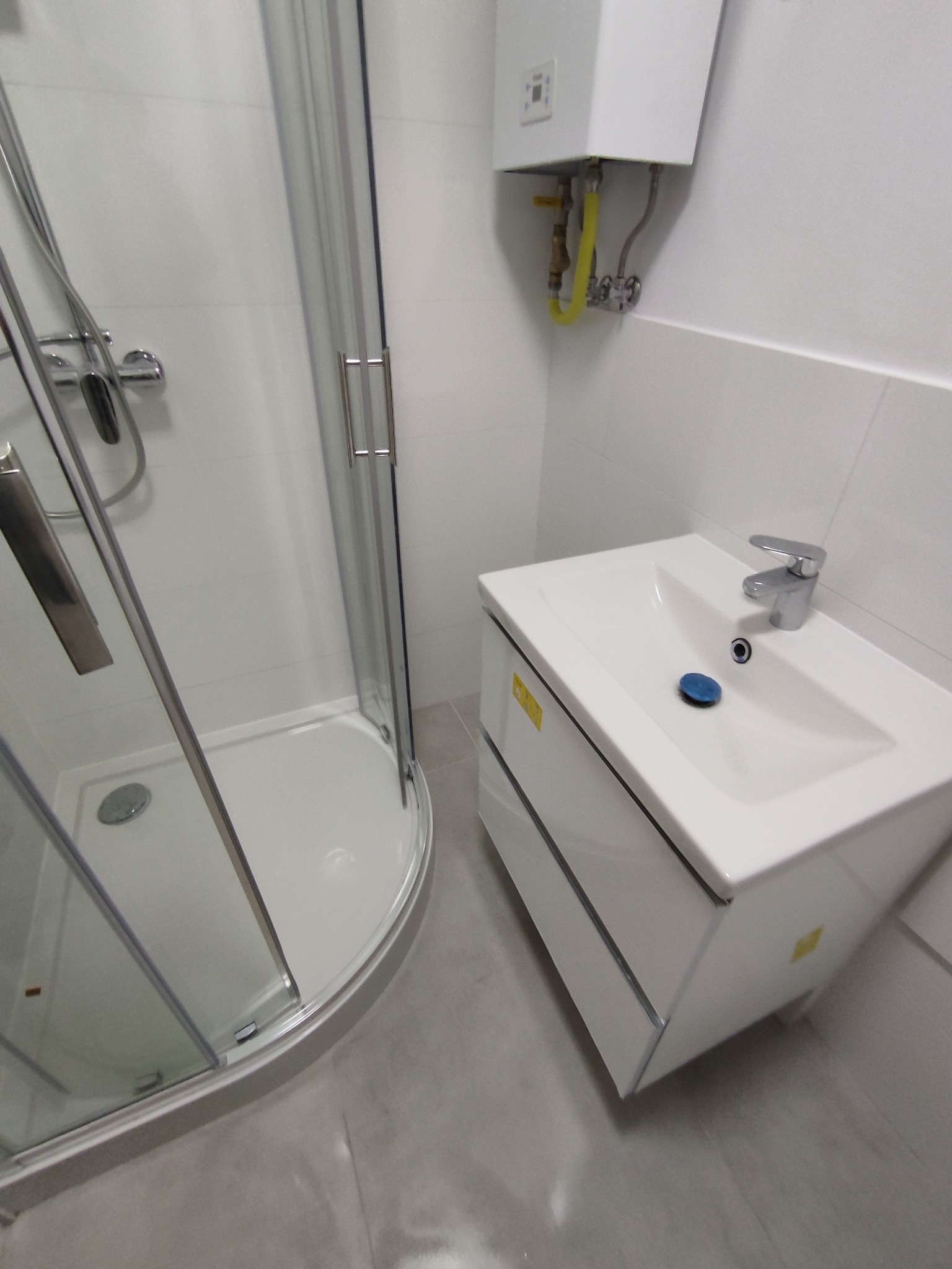 Nowoczesna łazienka z narożnym prysznicem z przezroczystego szkła i białą szafką pod umywalkę z chromowaną baterią, na ścianie biały piecyk gazowy.