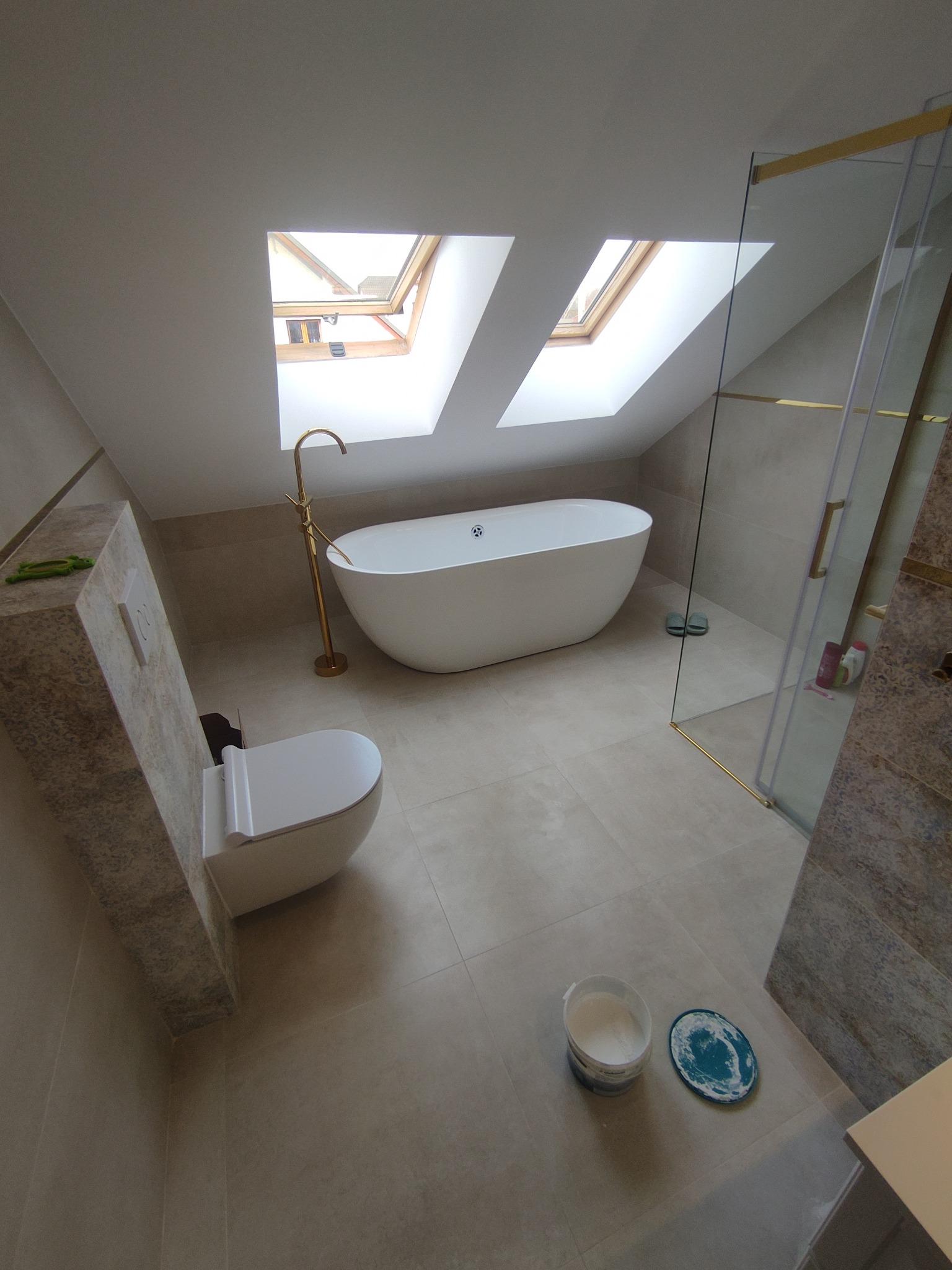 Jasna łazienka na poddaszu z wolnostojącą wanną, złotą armaturą, kabiną prysznicową ze złotymi detalami, toaletą i dwoma oknami dachowymi.