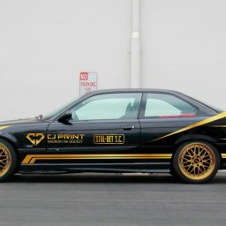 SOLO STUDIO Studio Graficzne Marcin Mikołajczyk - Czarny samochód BMW E36 z złotymi felgami i naklejonymi złotymi paskami oraz logo firm CJ Print i Stal-Bet S.C. na białym tle ściany.