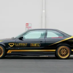 Czarny samochód BMW E36 z złotymi felgami i naklejonymi złotymi paskami oraz logo firm CJ Print i Stal-Bet S.C. na białym tle ściany.