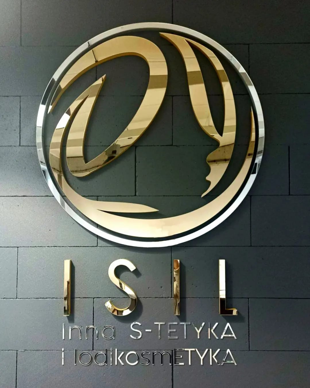 Złote logo ISIL z profilem twarzy w okręgu na szarej ścianie z cegieł, podświetlone, z napisem 'Inna S-TETYKA i lodikosmETYKA'.