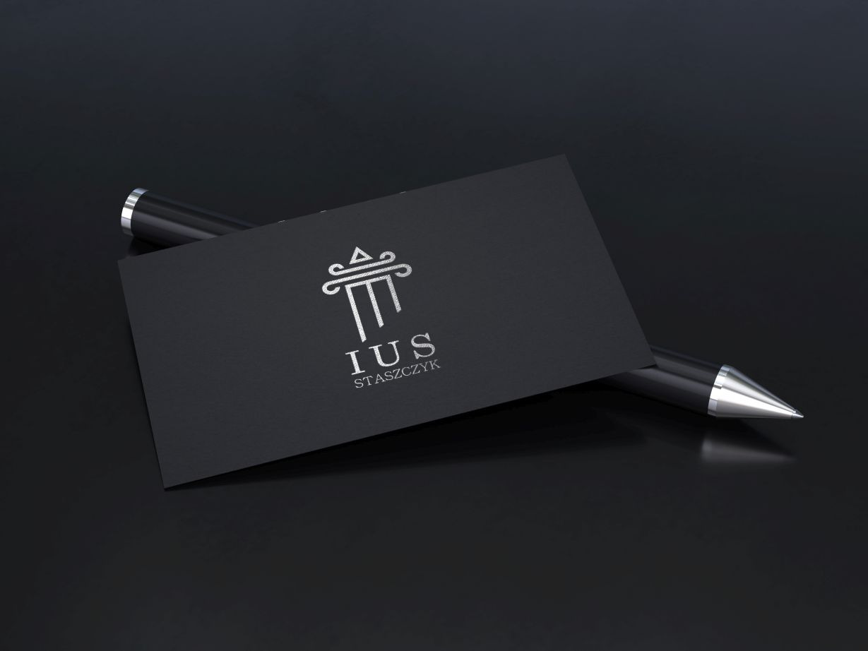 Elegancka wizytówka firmy prawniczej IUS Staszczyk z srebrnym logo kolumny na czarnym tle, obok leży czarny długopis.